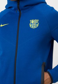 Μπλε φούτερ Nike με μαύρο φερμουάρ, κίτρινο λογότυπο Barcelona και αντίθετη ραφή κατά μήκος των ραφών. Μαλακό, υφασμάτινο ύφασμα.