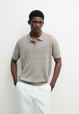 Jonge man die een beige polo met korte mouwen en een witte broek draagt, staand met handen in de zakken tegen een effen witte achtergrond.