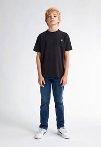 Černé bavlněné tričko s kulatým výstřihem a kruhovým logem, kombinováno s modrými džínami a bílými teniskami. Jednobarevné pozadí.