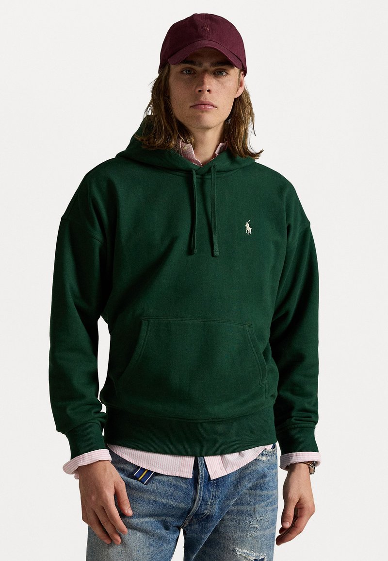 Grøn hættetrøje med en frontlomme og snor, parret med en pink og hvid stribet skjorte, slidte blå jeans og en bordeaux cap.