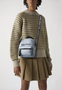 Šedý batoh z umelého kože s predným vreckom na zips, nastaviteľným popruhom a logom. Model má na sebe pruhovanú mikinu a plisovanú sukňu.