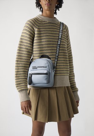 Mochila de piel sintética gris con un bolsillo frontal con cremallera, correa ajustable y marca. El modelo lleva un suéter a rayas y una falda con pliegues.