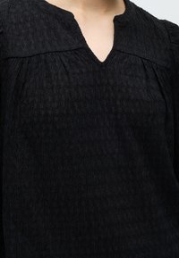 Черна риза с текстура и малък V-образен разрез на деколтето, носена от човек, видим от брадичката до гърдите.