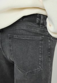 Jean en denim gris foncé avec une poche arrière, mise en avant. La texture est lisse, et une étiquette en cuir noir est visible à la taille.