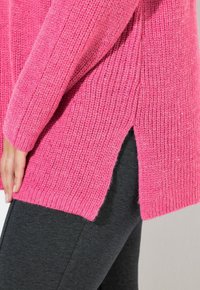 Rosa gestrickter Pullover mit Rippstrickmuster und Seitenschlitzen, getragen über dunkelgrauen Hosen. Der Pullover hat eine lockere, entspannte Passform.