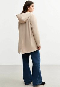Beige vattentät jacka med huva, med rak skärning och ventil bak. Matchad med vida denimbyxor i en mörkblå färg.