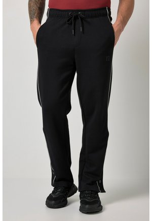 Man draagt zwarte trainingsbroek met witte zijstrepen, elastische tailleband en drukknoopsluiting bij de enkels, gecombineerd met zwarte sneakers.