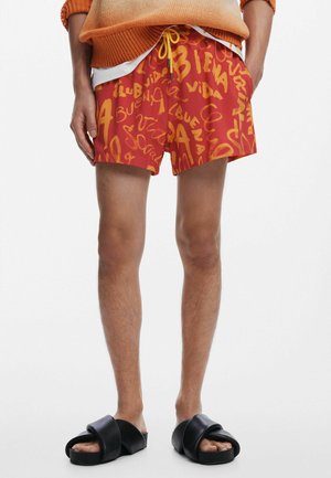 Red swim shorts featuring bold, yellow script text. Includes an elastic waistband with a drawstring. Worn with black slide sandals.