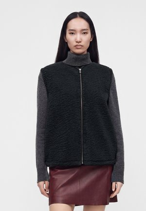 Frau mit langen dunklen Haaren trägt eine schwarze Fleeceweste mit Reißverschluss über einem grauen Rollkragenpullover und einem dunkelroten Lederrock.