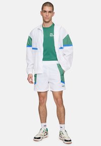 Rompevientos blanco con acentos en verde y azul, camiseta verde, pantalones cortos blancos con detalles verdes y zapatillas deportivas. Presenta un diseño deportivo y casual.