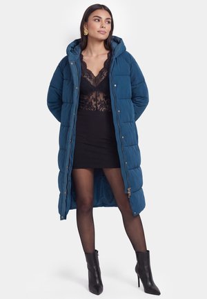 Frau trägt einen langen blauen Daunenmantel über einem schwarzen Spitzen-Top und Rock mit transparenten Strumpfhosen und schwarzen hochhackigen Stiefeletten.