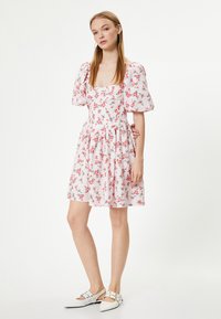 Bloemenjurk met pofmouwen, met een lichtroze basis en kleine rode bloemen, een geraffineerde taille en een uitlopende rok. Gecombineerd met witte schoenen.