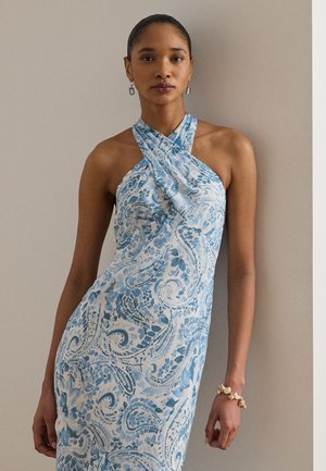 Γυναίκα με μαλλιά τραβηγμένα πίσω, που φοράει μπλε και λευκό φόρεμα χιαστί με σχέδιο πασλέι, βραχιόλι με πέρλες και κρεμαστά σκουλαρίκια, που στέκεται μπροστά σε έναν απλό τοίχο.