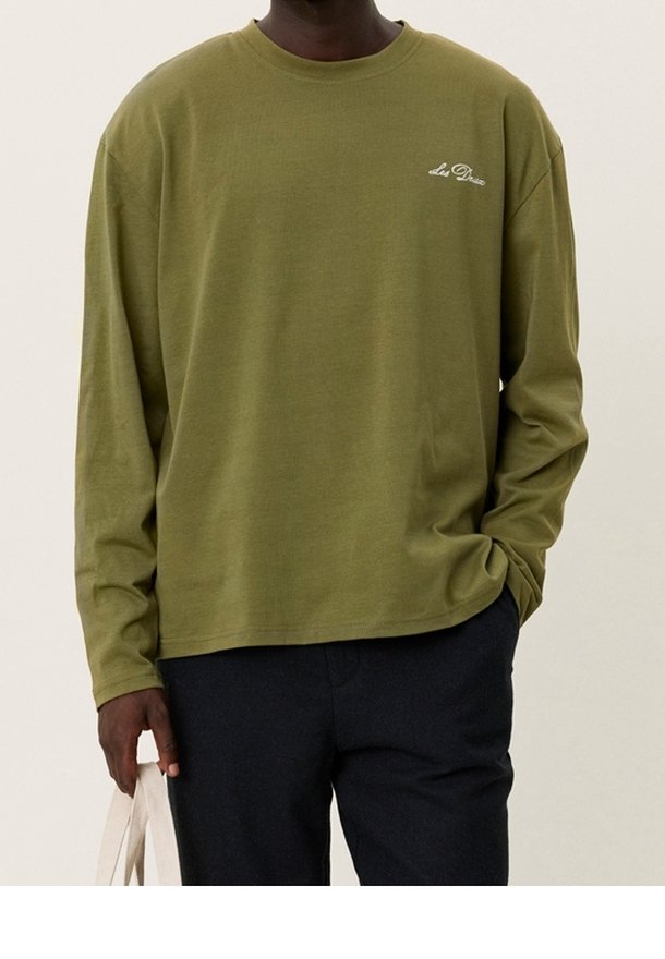 Olivenfarbendes Langarmshirt aus Baumwolle mit Rundhalsausschnitt und kleinem weißen Logo auf der Brust. Kombiniert mit dunklen Hosen.