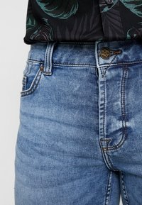 Jeans in denim blu chiaro con una texture leggermente sbiadita, dotati di una chiusura con bottoni e di un classico design a cinque tasche. Cuciture decorative visibili.