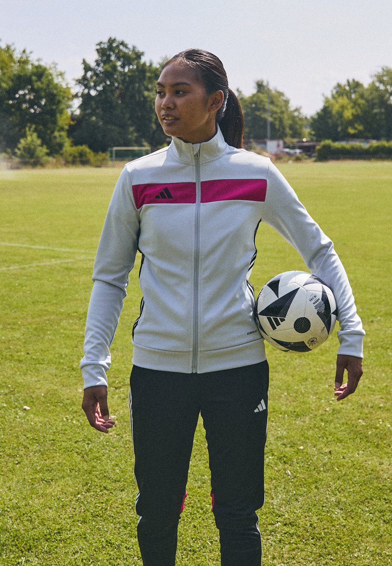 Kurtka sportowa Adidas w kolorze białym z różowym paskiem, czarne spodnie dresowe, trzymająca piłkę nożną w czarno-białe wzory na zielonej trawie.