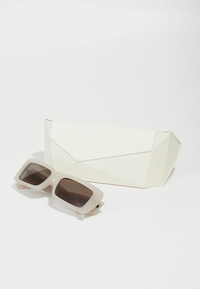 Lunettes de soleil blanches rectangulaires avec des verres foncés reposant à côté d'un étui à lunettes de soleil géométrique blanc assorti sur une surface blanche.