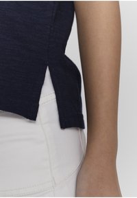 Námořnická modrá košile s krátkým rukávem a bočními rozparky, vyrobená z texturované látky, nošená přes bílé kalhoty. Detailní záběr zaměřený na košili a detaily okrajů.