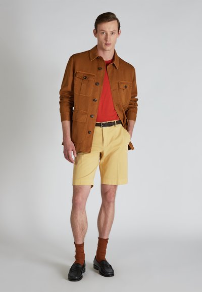 Chaqueta marrón de botones con dos bolsillos en el pecho, camiseta roja, pantalones cortos amarillos, calcetines marrones y mocasines negros sobre un fondo claro.