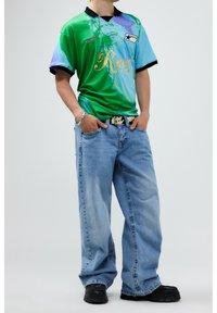 Polo gráfico verde y azul con cuello negro, texto dorado "Rave", combinado con jeans holgados azul claro y zapatos negros de suela gruesa.