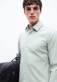 Νεαρός άνδρας με κοντά σγουρά μαλλιά, που φοράει ανοιχτό πράσινο πουκάμισο με κουμπιά, κρατώντας μαύρη δερμάτινη τσάντα σε λευκό φόντο.