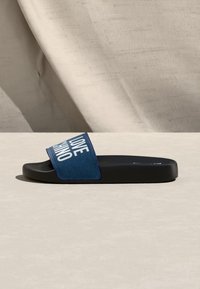 Μονή μαύρη σαγιονάρα με μπλε υφασμάτινο λουράκι που φέρει λευκό κείμενο "LOVE MOSCHINO" σε ανοιχτή επιφάνεια με μαλακές σκιές.