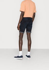 Shorts de mezclilla oscura con dobladillo vuelto, combinados con una camiseta ligera de melocotón y zapatillas de lona navy. Calcetines blancos visibles por encima de las zapatillas.