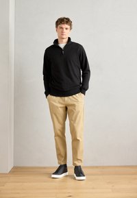 Black zip-up sweatshirt with high collar, beige chinos, and black sneakers with white soles, standing against a light gray background.