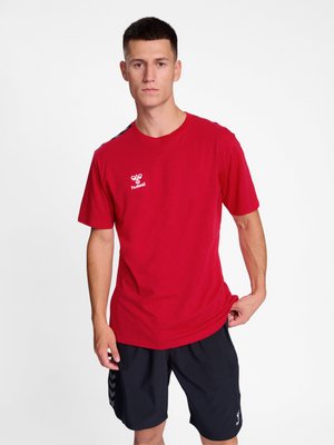 Giovane uomo con capelli corti che indossa una maglietta rossa Hummel e pantaloncini sportivi neri su uno sfondo bianco uniforme.