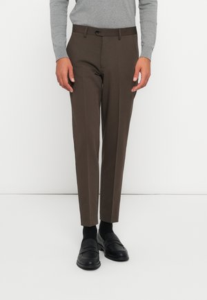 Pantalons marron sur mesure en tissu lisse, avec une coupe slim, un devant plat et une fermeture à un bouton. Associés avec des mocassins noirs.