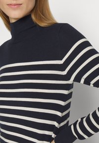 Nainen, joka pitää yllään tummansinistä poolopaitaa, jossa on valkoisia vaakaraitoja, käsi koukistetuna vyötärön kohdalla yksinkertaisen taustan edessä.