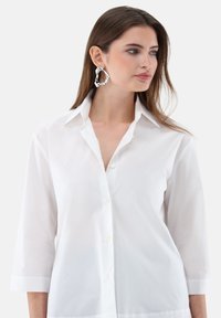 White button-up shirt with three-quarter sleeves, made of smooth fabric, featuring a classic collar and subtle sheen. Large, silver hoop earrings.