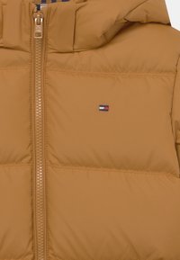 Brązowa kurtka puchowa z wysokim kołnierzem i zapięciem na zamek. Posiada małe logo na klatce piersiowej oraz gładką, teksturowaną zewnętrzną tkaninę.
