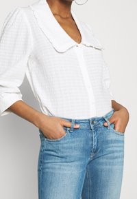 Kvinna som bär en vit texturerad blus med en stor volangkrage och ljusblå högmidjade jeans, händerna i fickorna, synliga guldringar i  öronen.