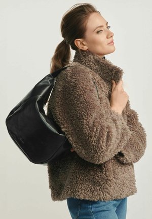 Czarna skórzana torba na ramię o gładkiej teksturze i zaokrąglonym wzorze. Modelka nosi puszysty jasnobrązowy płaszcz i niebieskie dżinsy, prezentując swobodny styl.