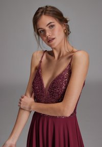 Joven mujer con vestido burdeos sin mangas, de escote en V profundo y con lentejuelas, con pendientes de aro dorados sobre un fondo gris liso.