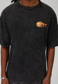 T-shirt oversized preta em um tecido texturizado e desbotado. Apresenta uma graphic de um sol sorridente e vibrante com chamas no lado esquerdo do peito.