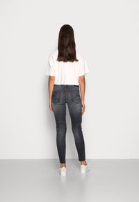 Žena s dlouhými hnědými vlasy, oblečená v bílém crop topu, tmavě šedých úzkých džínách a bílých teniskách, stojí čelem k bílé zdi.