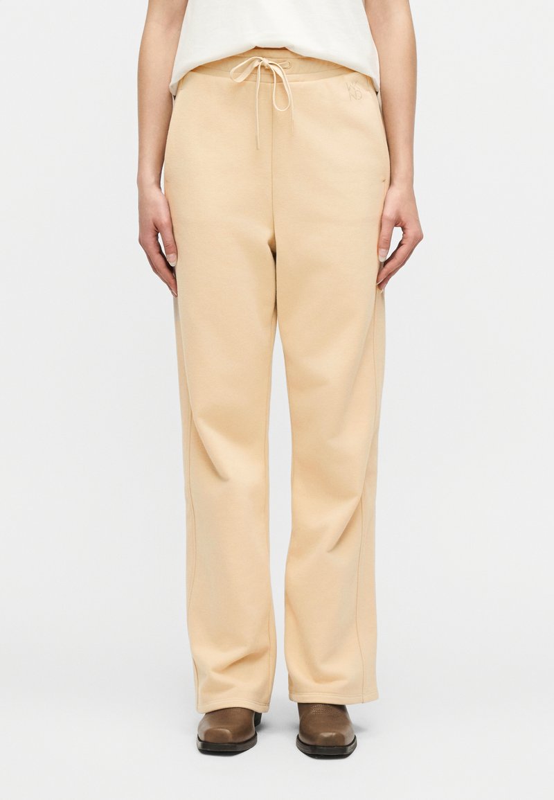 Άτομο που φοράει φαρδιά μπεζ παντε λόνια φόρμας με κορδόνι στη μέση και καφέ μπότες αστραγάλου, στέκεται μπροστά σε απλό φόντο.