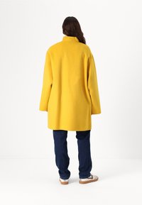 Cappotto di lana giallo con colletto alto, maniche lunghe e taglio dritto, abbinato a jeans blu scuro e sneakers chiare.