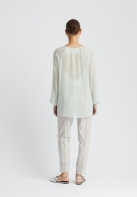 Helles grün, durchscheinendes Blusenshirt mit langen Ärmeln, einem runden Ausschnitt und Falten im Rücken, kombiniert mit hellbeigen, schmal zulaufenden Hosen und Sandalen mit silbernen Akzenten.