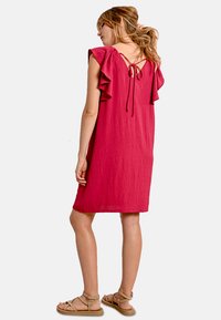 Abito a maniche corte arruffato in un vivace rosa, realizzato in tessuto testurizzato. Presenta una chiusura cravatta sul retro e una vestibilità rilassata, che termina sopra il ginocchio.