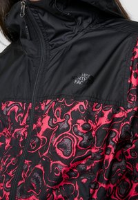 Czarna i różowa kurtka w wzory z zamkiem błyskawicznym, wysokim kołnierzem i detalem z logo. Tkanina ma gładką teksturę i sportowy design.