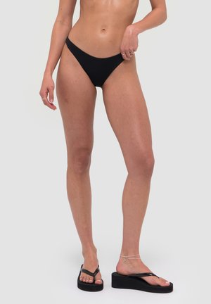 Kobieta ubrana w czarne spodnie od bikini i czarne klapki na platformie, stojąca na jednolitym jasnoszarym tle.