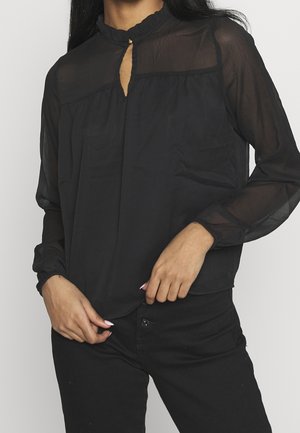 Vrouw draagt een doorschijnende zwarte blouse met lange mouwen en een kleine oogvormige halslijn, en zwarte broek, met handen die de zoom van de blouse licht vasthouden.