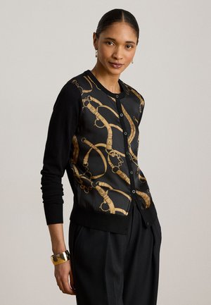 Femme portant un cardigan noir boutonné avec des motifs dorés de chaînes et ceintures style équestre, un pantalon noir, un bracelet doré et de petites boucles d'oreilles créoles.