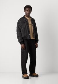 Black bomber jacket with snap buttons, beige sweatshirt with a logo, black cargo pants, and brown slip-on shoes with thick soles.
