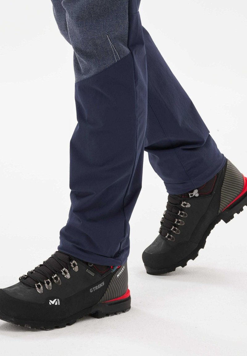 Bottes de randonnée noires avec œillets en métal et un talon texturé gris et rouge, associées à un pantalon de plein air bleu marine avec une partie supérieure à motifs.