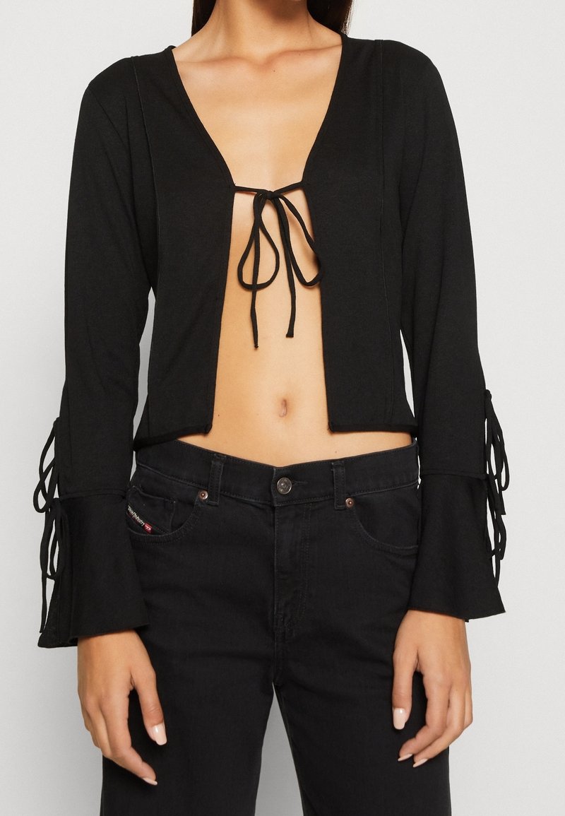 Femme portant un haut noir court à manches longues noué à la poitrine et un jean noir taille haute.
