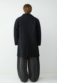 Langer schwarzer Mantel mit geradem Schnitt, weiten Ärmeln und Kragen. Kombiniert mit locker sitzenden, dunkelgrauen Hosen. Glatte Textur.